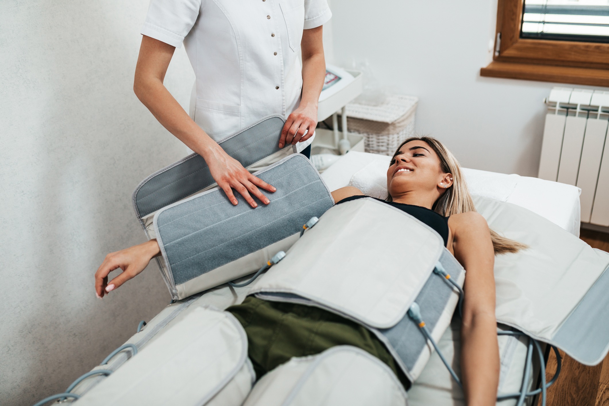 A woman is lying on a bed undergoing a body contouring or massage treatment. She is smiling and covered with gray wraps on her arms and torso. A professional stands beside her, adjusting the equipment.
