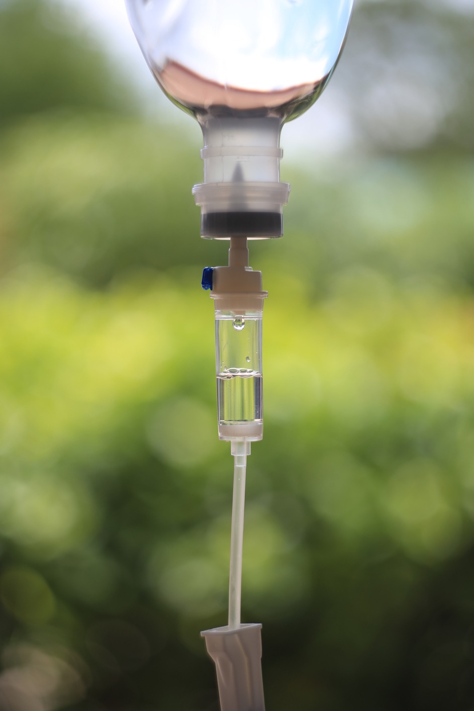 Close-up of an intravenous (IV) drip with clear fluid inside the chamber, against a blurred green background.