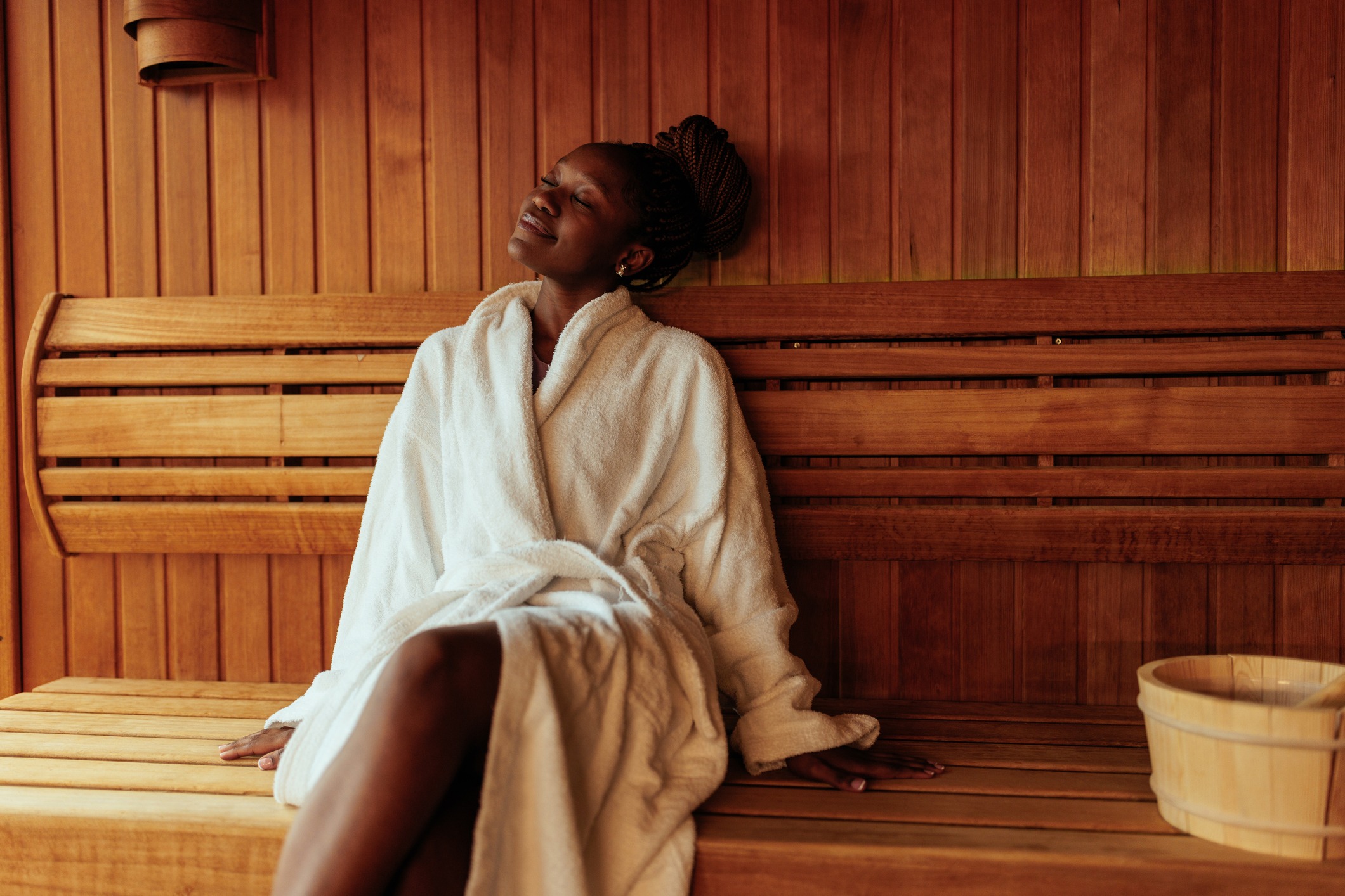 A woman wearing a white bathrobe relaxes on a wooden bench inside a sauna, eyes closed and smiling peacefully, with a wooden bucket nearby.