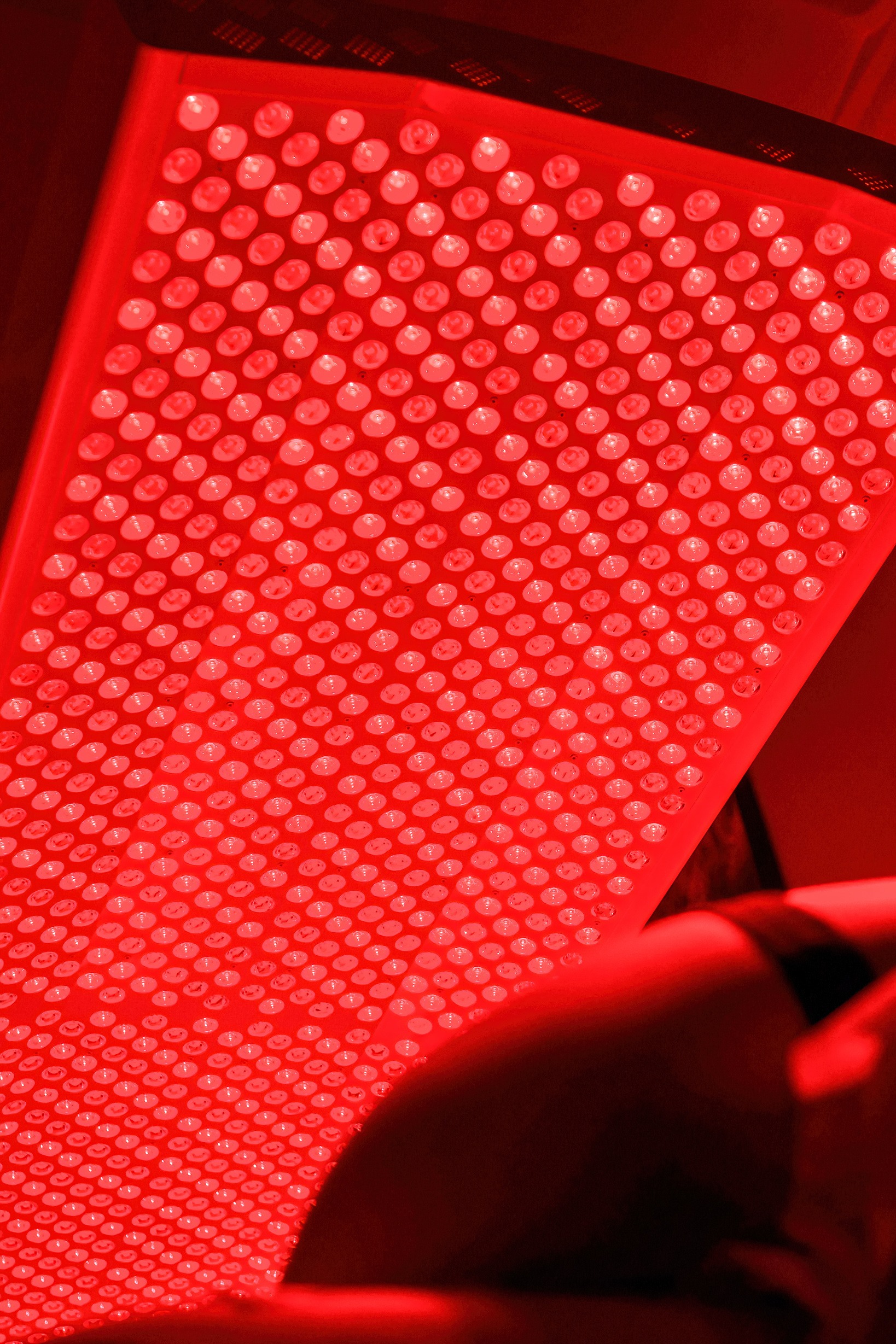 A close-up, angled view of a red LED light therapy panel glowing brightly, with a blurred part of a person’s shoulder and back visible in the foreground.