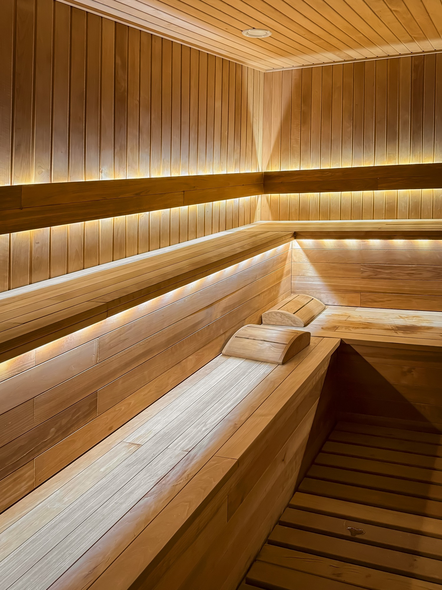 A modern wooden sauna interior with warm, ambient lighting, tiered bench seating, and wooden headrests, creating a relaxing and inviting atmosphere.